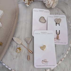 Puede incluir: Tres alfileres de corazón dorados con detalles de purpurina blanca. Los alfileres se muestran en tarjetas blancas con el texto "Handmade by TERESA LOVES CRAFTS" y la dirección del sitio web "TeresaLovesCrafts.etsy.com".