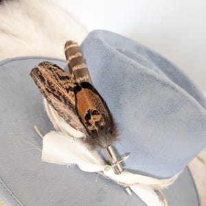 Può includere: Un cappello fedora azzurro con un accento decorativo di piume e tessuto. Il cappello ha un'ampia tesa e una morbida texture simile alla pelle scamosciata. Le piume sono marroni, nere e arancioni.