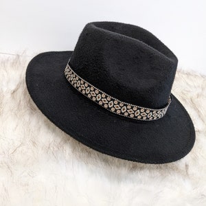 Op de afbeelding: Zwarte fedora-hoed met een decoratieve band met een herhalend patroon van kleine, ruitvormige ontwerpen in bruin- en crèmetinten. De hoed heeft een brede rand en een licht gestructureerd oppervlak.