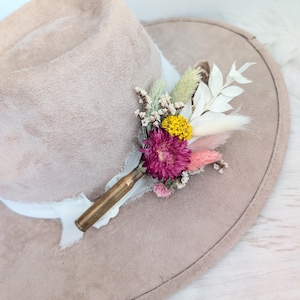 Può includere: Un cappello fedora in camoscio beige con un nastro bianco, adornato con una composizione floreale. La composizione comprende un fiore rosa intenso, fiori gialli e varie erbe e foglie secche, attaccate a un bossolo di proiettile in ottone.