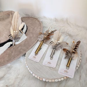 Può includere: Un cappello da cowboy beige con una fascia a righe bianche e nere è adornato da un accento di piume e fiori secchi. Tre spille a bossolo con composizioni di piume e fiori secchi sono esposte su un vassoio di legno.