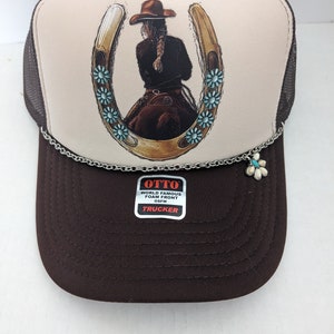 May include: A brown and white trucker hat with a horseshoe and a woman riding a horse design. The hat has a silver chain with a flower charm. The hat has a label that says "OTTO WORLD FAMOUS FOAM FRONT OS FM TRUCKER".