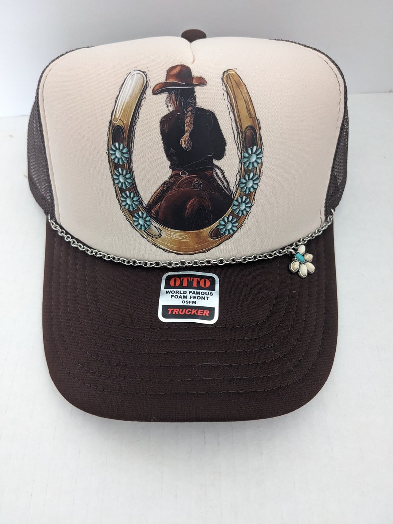 May include: A brown and white trucker hat with a horseshoe and a woman riding a horse design. The hat has a silver chain with a flower charm. The hat has a label that says "OTTO WORLD FAMOUS FOAM FRONT OS FM TRUCKER".