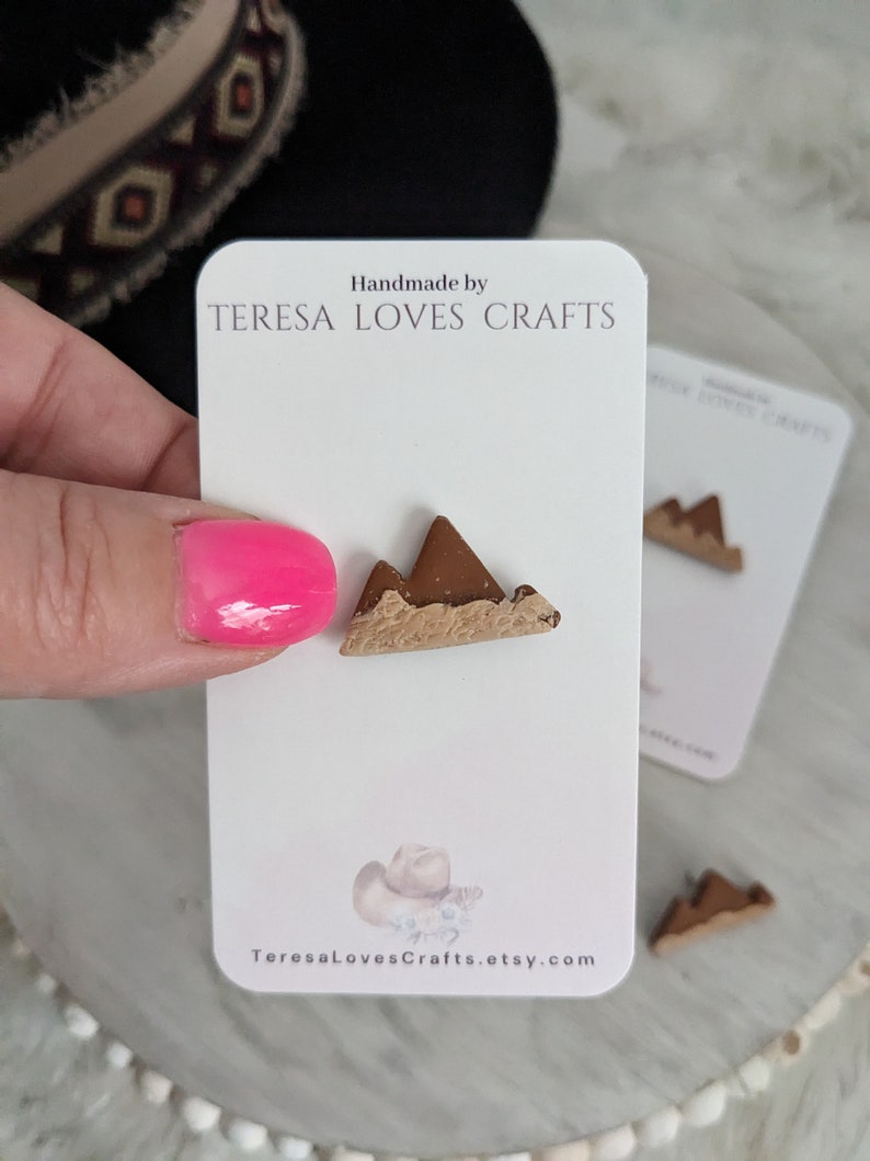 May include: Handmade by TERESA LOVES CRAFTS, a pair of brown mountain-shaped polymer clay earrings. The earrings are on a white card with the shop's name and website address.