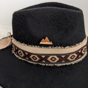 May include: Black felt fedora hat with a brown leather mountain design on the crown. The hat has a tan band with a geometric pattern in black, red, and beige.