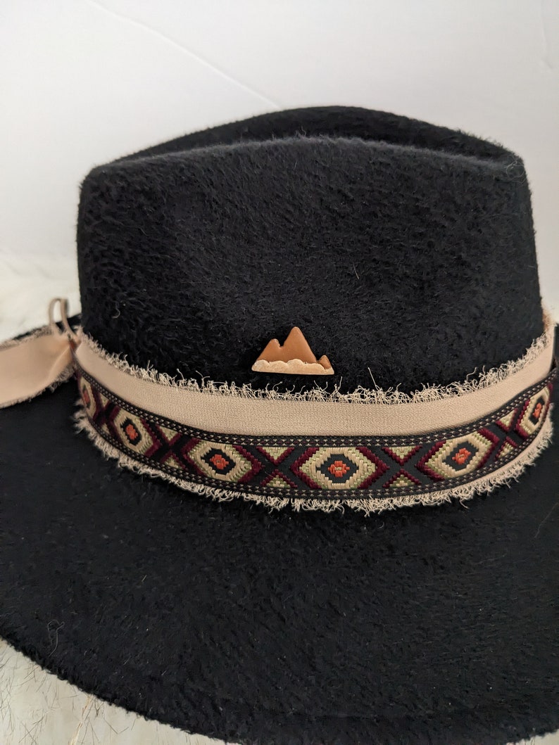 May include: Black felt fedora hat with a brown leather mountain design on the crown. The hat has a tan band with a geometric pattern in black, red, and beige.