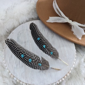 May include: Two black and white feathers with turquoise accents on a white wooden base with a brown hat in the background.