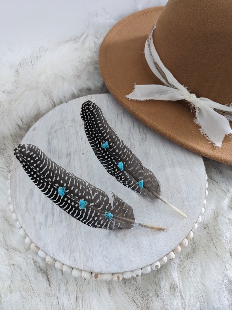 May include: Two black and white feathers with turquoise accents on a white wooden base with a brown hat in the background.