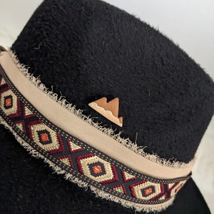 May include: Black felt fedora hat with a tan band featuring a geometric pattern. The band has a frayed edge. A small brown leather mountain pin is attached to the hat.