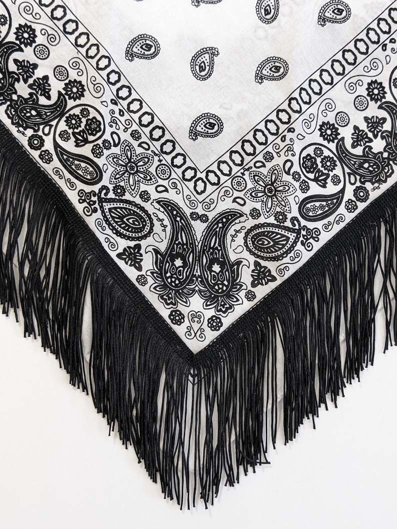 May include: A white triangular bandana featuring a black paisley and floral pattern throughout the fabric. The edges of the bandana are finished with long, thin black fringe trim that hangs down from the two sides of the triangular point.