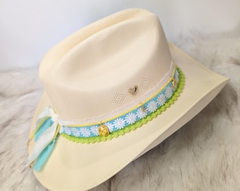 May include: Cream-coloured cowboy hat with a decorative band featuring white daisy flowers, blue and yellow accents, and gold charms. The brim is trimmed with green scalloped lace. A blue and white ribbon with feathers is tied on the side.