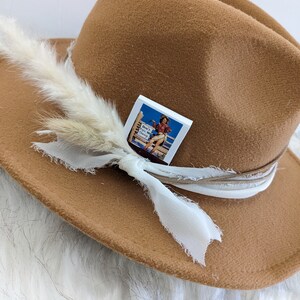 Pu&ograve; includere: Un cappello fedora in feltro marrone con un nastro bianco e un piccolo spillo quadrato bianco con un'immagine vintage di una donna a cavallo. Lo spillo dice "Don't Get In My Way Honey".