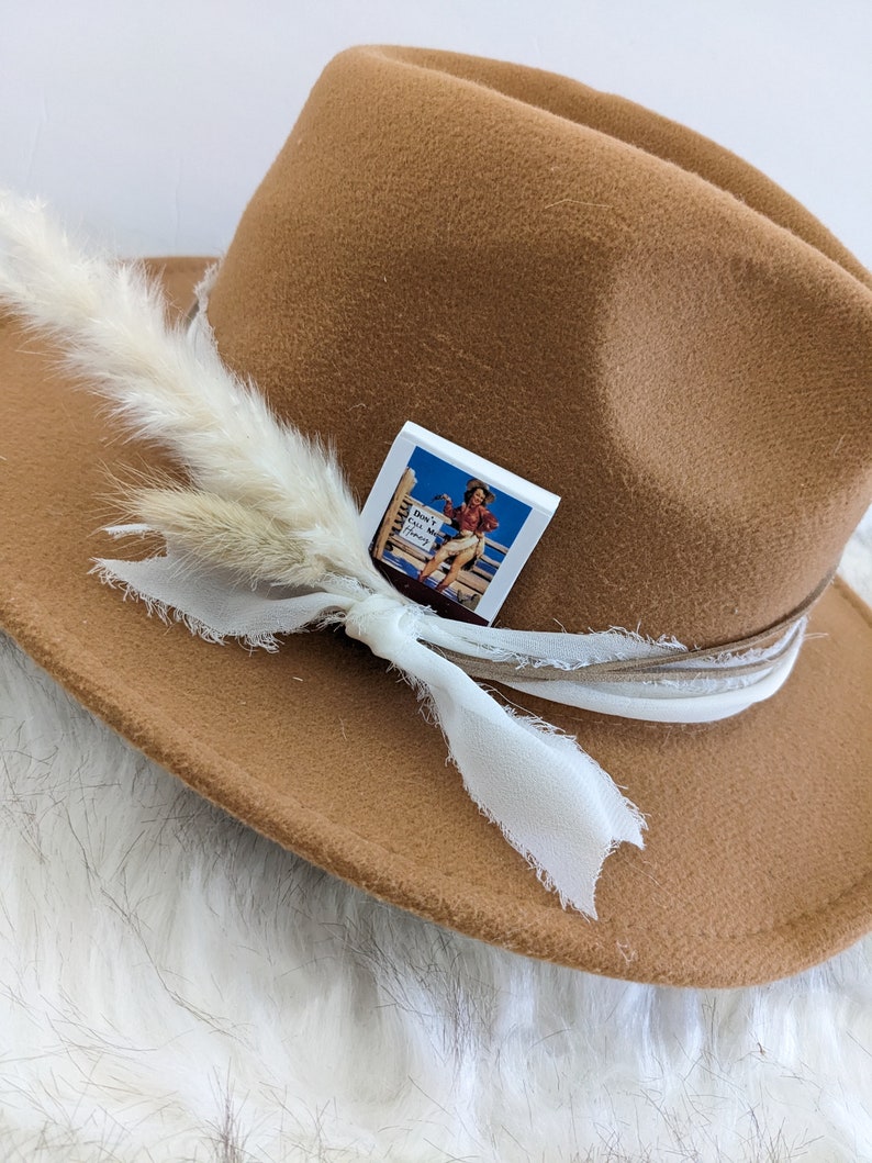 Pu&ograve; includere: Un cappello fedora in feltro marrone con un nastro bianco e un piccolo spillo quadrato bianco con un'immagine vintage di una donna a cavallo. Lo spillo dice "Don't Get In My Way Honey".