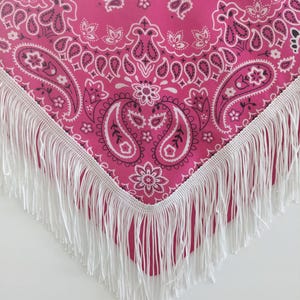 May include: A bright pink bandana featuring a traditional white and black paisley pattern. The triangular fabric is finished along the two bottom edges with a long, straight white fringe trim that hangs down from the hem of the textile.