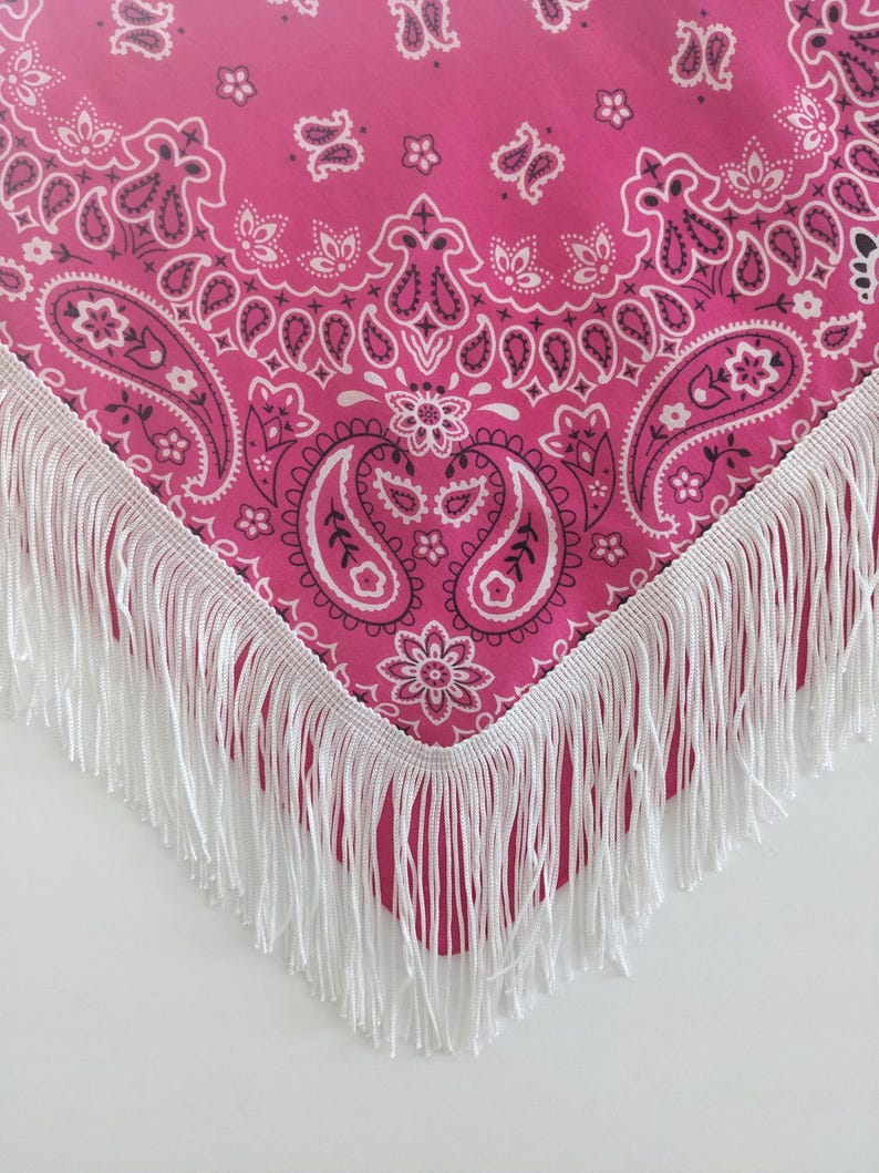 May include: A bright pink bandana featuring a traditional white and black paisley pattern. The triangular fabric is finished along the two bottom edges with a long, straight white fringe trim that hangs down from the hem of the textile.