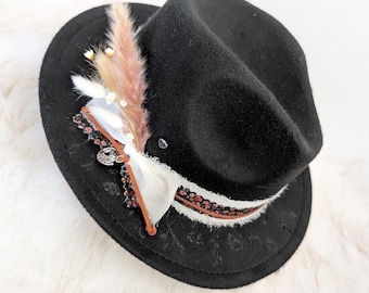May include: A black fedora hat with a decorative band and floral embellishments. The hat features a white ribbon, brown leather trim, and a cluster of dried flowers and feathers. The brim has a subtle floral pattern.
