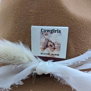 Pu&ograve; includere: Un cappello da cowboy in feltro marrone con un nastro bianco e una piuma bianca. Un piccolo cartellino quadrato bianco &egrave; attaccato al nastro con il testo "Cowgirls Kick Ass" e un'immagine in bianco e nero di una donna a cavallo.
