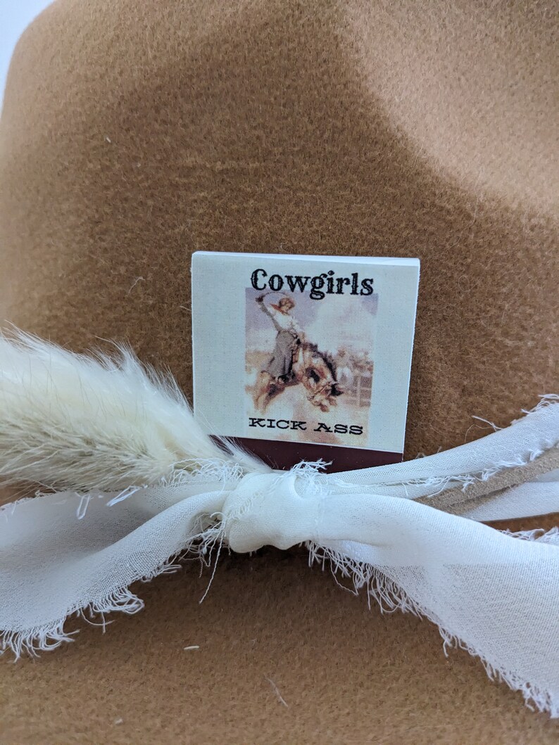 Pu&ograve; includere: Un cappello da cowboy in feltro marrone con un nastro bianco e una piuma bianca. Un piccolo cartellino quadrato bianco &egrave; attaccato al nastro con il testo "Cowgirls Kick Ass" e un'immagine in bianco e nero di una donna a cavallo.