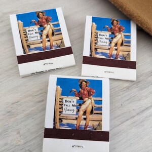 Pu&ograve; includere: Tre fiammiferi con un'illustrazione in stile vintage di una donna con un cappello da cowboy e un abbigliamento da cowgirl. La donna &egrave; in piedi davanti a una recinzione di legno con un cartello che dice "Non chiamarmi tesoro".