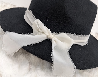May include: A black felt fedora hat with a white ribbon tied in a bow. The ribbon has a frayed edge.