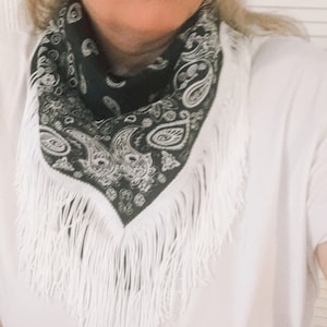 May include: A black bandana featuring a white paisley pattern and long white fringe trim along the edges. The triangular-folded fabric is worn around the neck over a plain white t-shirt, showcasing the intricate contrast of the monochrome design.