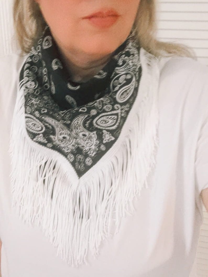 May include: A black bandana featuring a white paisley pattern and long white fringe trim along the edges. The triangular-folded fabric is worn around the neck over a plain white t-shirt, showcasing the intricate contrast of the monochrome design.