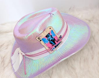 Könnte beinhalten: Schillernder Cowboyhut in Rosa- und Lilatönen, mit weißem Band und einer dekorativen Karte mit einer Pferdeillustration. Der Hut hat eine breite Krempe und einen Federakzent.