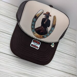 May include: A brown and beige trucker hat with a brown mesh back. The front of the hat features a graphic of a woman riding a horse with a horseshoe and turquoise flowers. The hat has a silver chain with a turquoise flower charm attached. The hat also has a label that says "OTTO WORLD FAMOUS FOAM FRONT 63FM TRUCKER".
