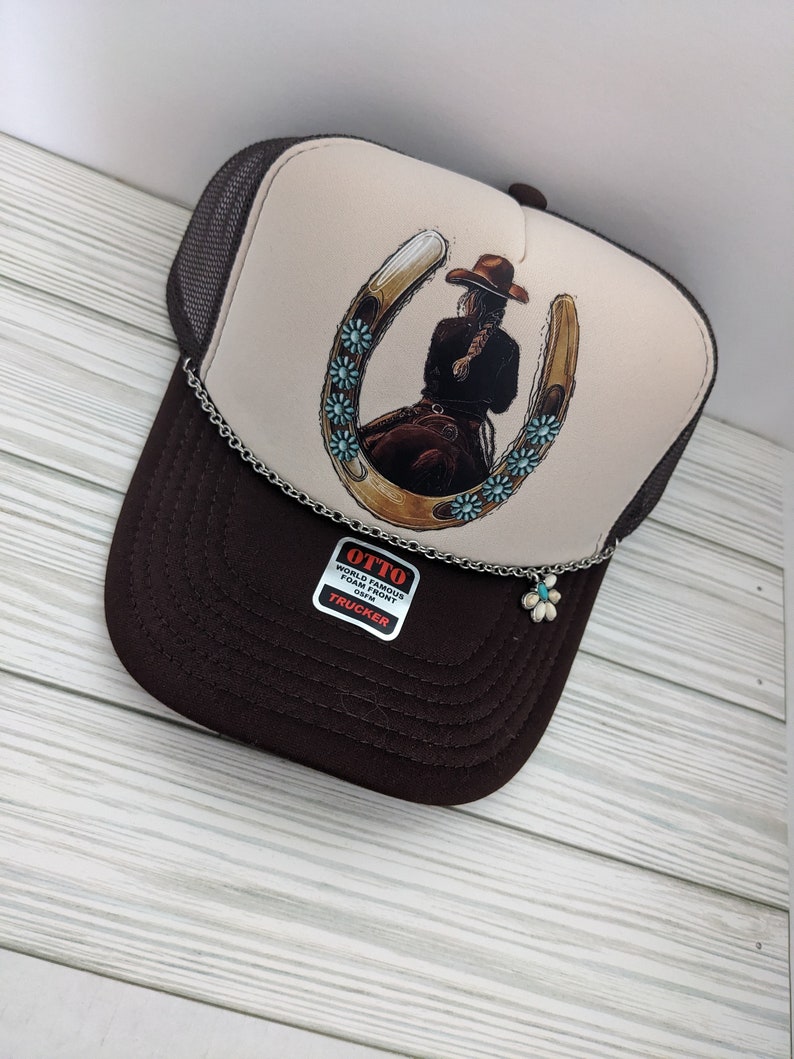 May include: A brown and beige trucker hat with a brown mesh back. The front of the hat features a graphic of a woman riding a horse with a horseshoe and turquoise flowers. The hat has a silver chain with a turquoise flower charm attached. The hat also has a label that says "OTTO WORLD FAMOUS FOAM FRONT 63FM TRUCKER".