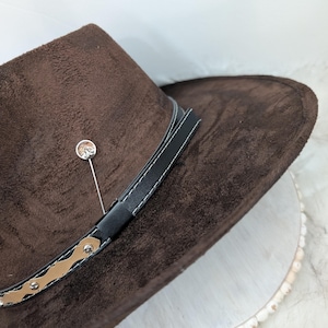 Puede incluir: Un sombrero vaquero de gamuza marrón con una banda de cuero negro y un adorno decorativo dorado. Un pequeño alfiler plateado está sujeto al sombrero.
