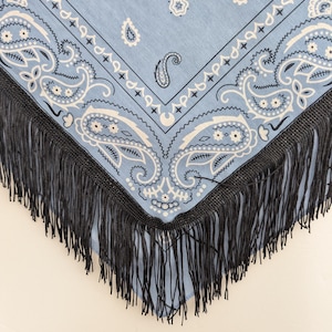 May include: A light blue bandana featuring a white paisley pattern with intricate floral and teardrop motifs. The fabric is bordered by a thick, dark black fringe trim that hangs from the edges, creating a distinct contrast against the blue.