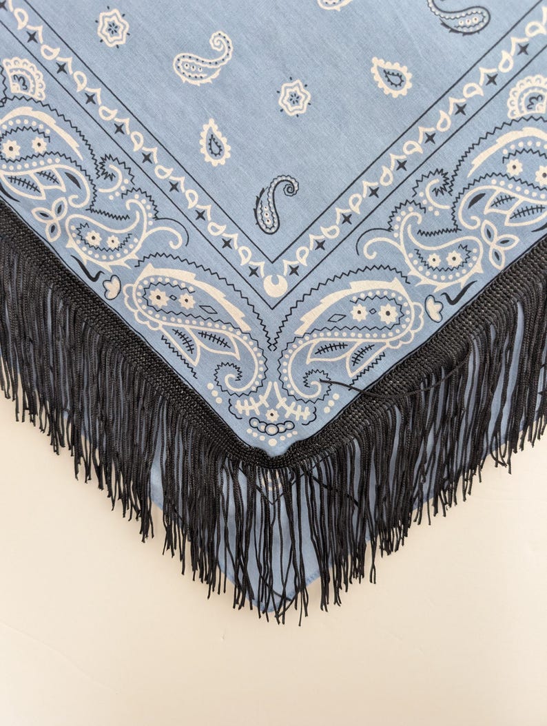 May include: A light blue bandana featuring a white paisley pattern with intricate floral and teardrop motifs. The fabric is bordered by a thick, dark black fringe trim that hangs from the edges, creating a distinct contrast against the blue.