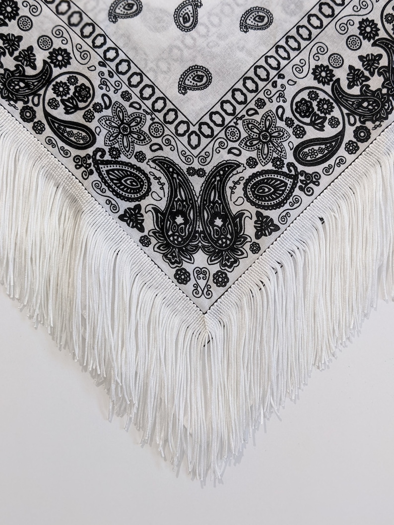 May include: A white triangular bandana featuring a black paisley print pattern. The edges of the fabric are adorned with long, straight white fringe trim that hangs down from the bottom point and sides of the textile accessory.