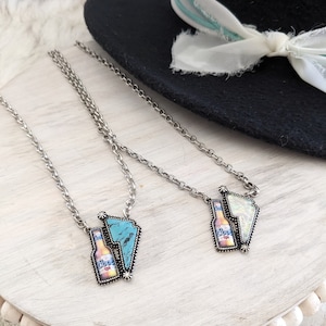 May include: Two silver chain necklaces with a broken beer bottle charm. The charms are made of metal and have a turquoise and white design. The necklaces are on a light wood surface.