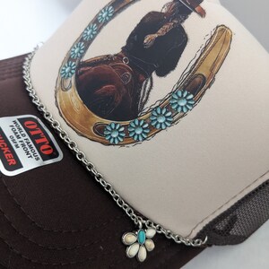 May include: A brown and beige trucker hat with a white horseshoe design and turquoise flowers. The hat has a silver chain with a turquoise and white flower charm.