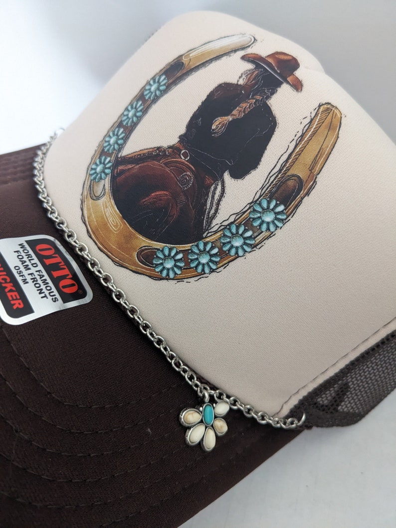 May include: A brown and beige trucker hat with a white horseshoe design and turquoise flowers. The hat has a silver chain with a turquoise and white flower charm.