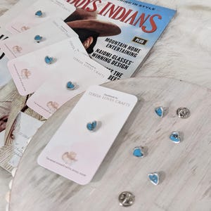 Op de afbeelding: Een set turquoise hartvormige oorbellen op een witte kaart met de tekst "Handmade by Teresa Loves Crafts". De oorbellen worden getoond op een wit oppervlak met een tijdschrift op de achtergrond.