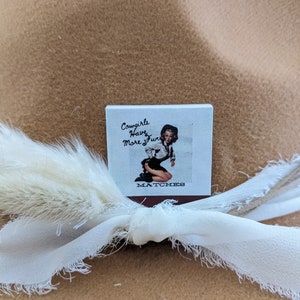 Pu&ograve; includere: Una scatola di fiammiferi con un design vintage che presenta una donna con un cappello da cowboy e il testo "Cowgirls Have More Fun".