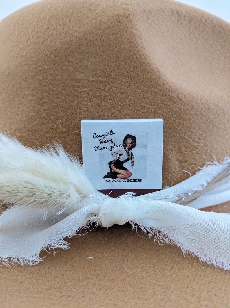 Pu&ograve; includere: Una scatola di fiammiferi con un design vintage che presenta una donna con un cappello da cowboy e il testo "Cowgirls Have More Fun".