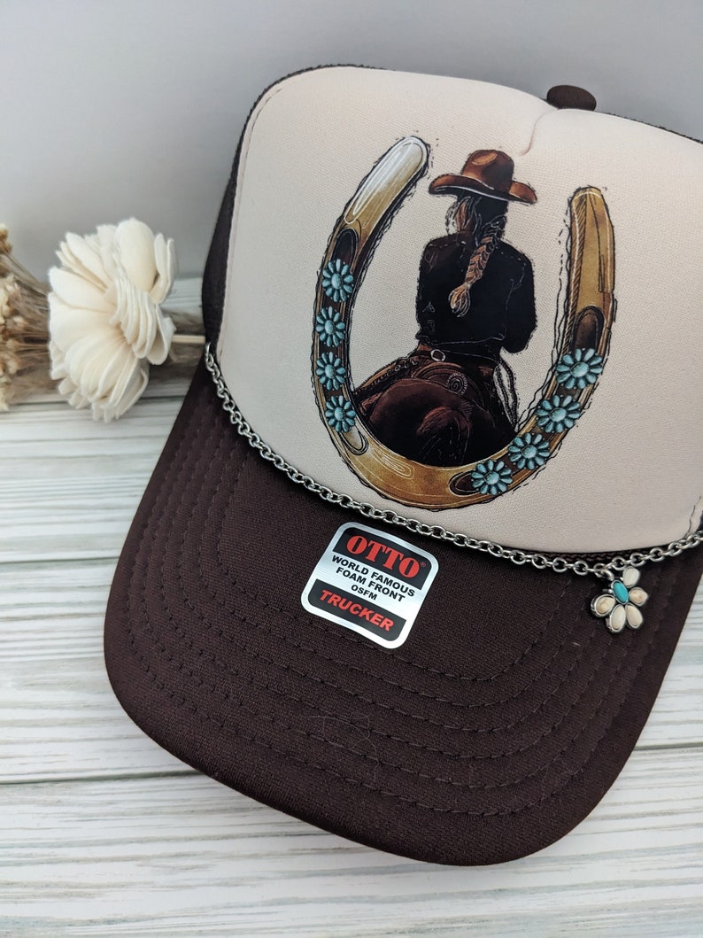 May include: A brown and white trucker hat with a horseshoe and a woman riding a horse design. The hat has a silver chain with a flower charm attached. The hat has a label that says "OTTO WORLD FAMOUS FOAM FRONT OSFM TRUCKER".