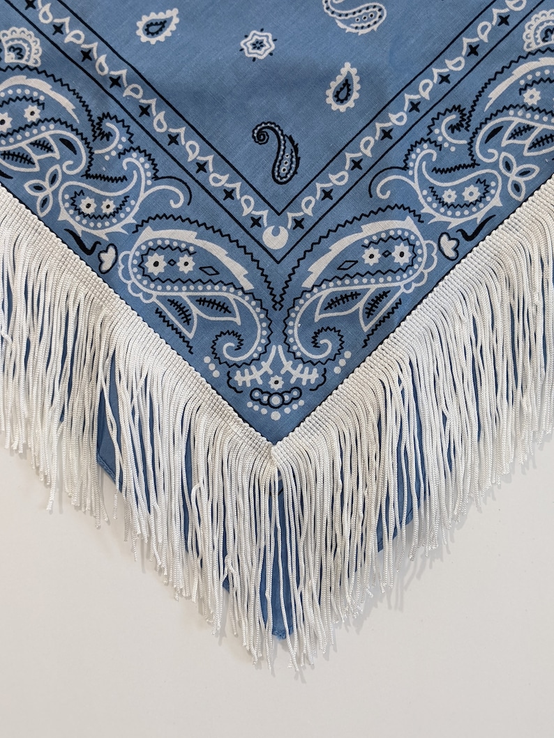 May include: A light blue bandana featuring a classic white and dark blue paisley pattern. The fabric is finished with a long, white fringe trim along the edges, creating a decorative border that hangs down from the triangular point of the cloth.