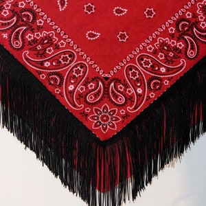 May include: A triangular red bandana featuring a black and white paisley pattern with floral motifs. The edges of the fabric are adorned with long, thin black fringe that hangs down from the bottom point and sides of the textile accessory.