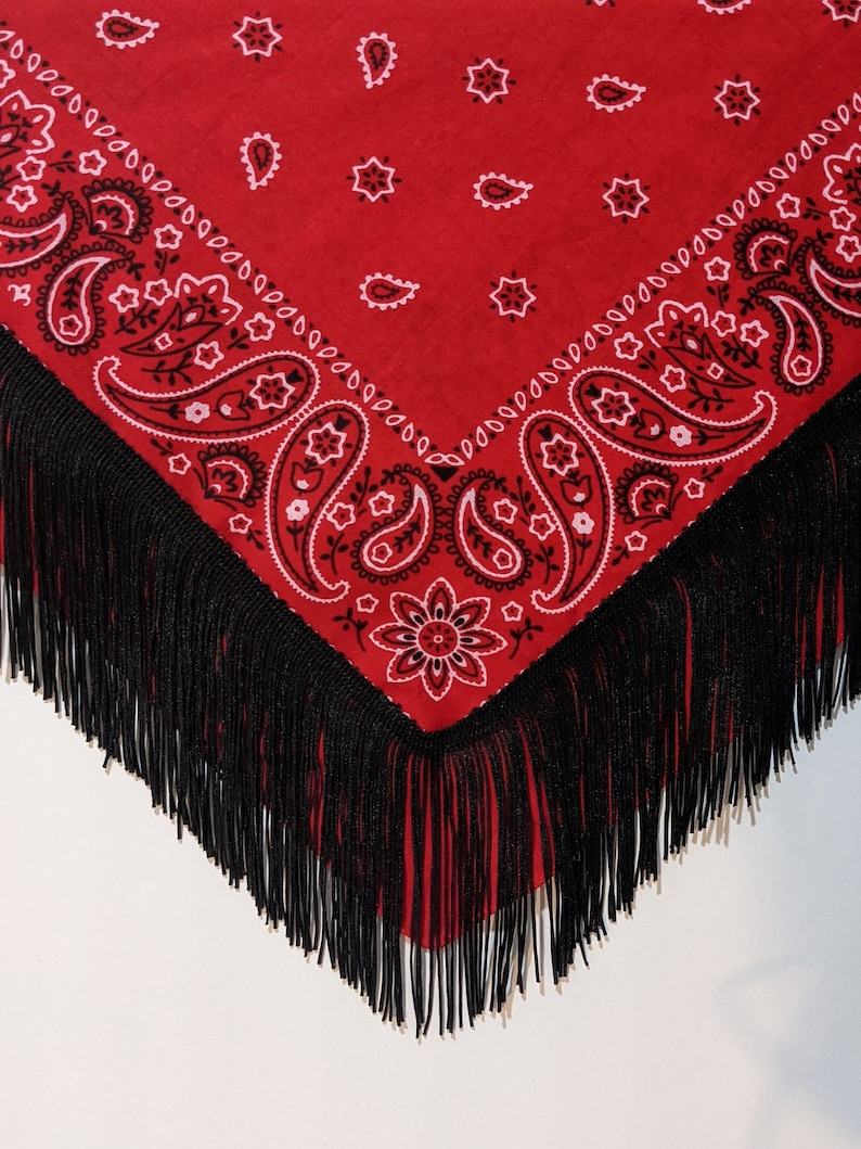 May include: A triangular red bandana featuring a black and white paisley pattern with floral motifs. The edges of the fabric are adorned with long, thin black fringe that hangs down from the bottom point and sides of the textile accessory.