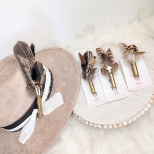 Può includere: Un cappello fedora beige con una fascia a righe bianche e nere, adornato con un gruppo di piume e un bossolo di proiettile in ottone. Tre spille con piume e bossoli di proiettile sono esposte su un vassoio bianco, ciascuna con disposizioni di piume uniche.