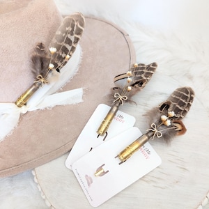 Può includere: Tre spille per cappello con piume con bossoli, piume e piccoli fiori bianchi. Le spille sono esposte su un cappello marrone chiaro e biglietti bianchi. Le carte hanno il testo "PinkMeCowboy.etsy.com".