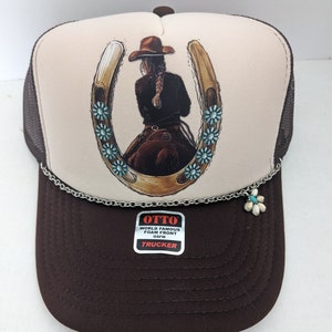 May include: A brown and white trucker hat with a horseshoe design featuring a woman riding a horse. The horseshoe is decorated with turquoise blue flowers. The hat has a silver chain with a flower charm attached. The hat has a label that reads "OTTO WORLD FAMOUS FOAM FRONT OSFM TRUCKER".
