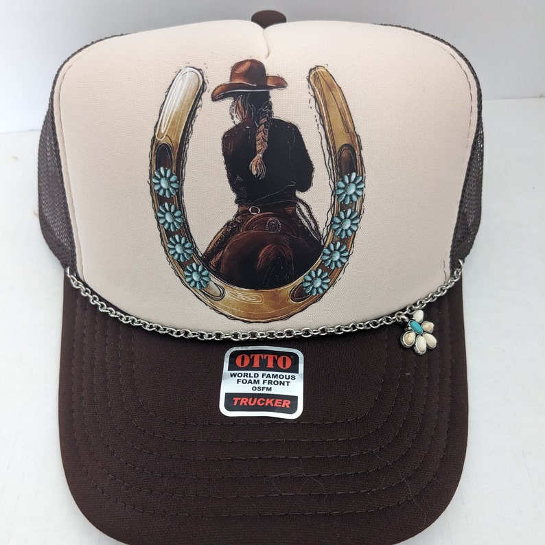 May include: A brown and white trucker hat with a horseshoe design featuring a woman riding a horse. The horseshoe is decorated with turquoise blue flowers. The hat has a silver chain with a flower charm attached. The hat has a label that reads "OTTO WORLD FAMOUS FOAM FRONT OSFM TRUCKER".