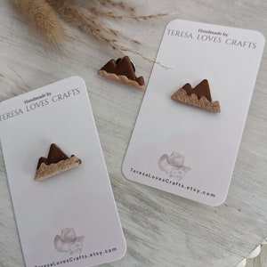May include: Two small brown and beige clay mountain-shaped pins on white cards with the text "Handmade by TERESA LOVES CRAFTS" and a website address.