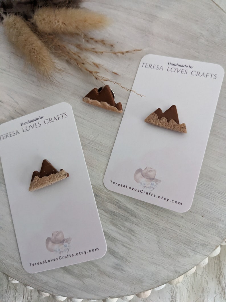 May include: Two small brown and beige clay mountain-shaped pins on white cards with the text "Handmade by TERESA LOVES CRAFTS" and a website address.