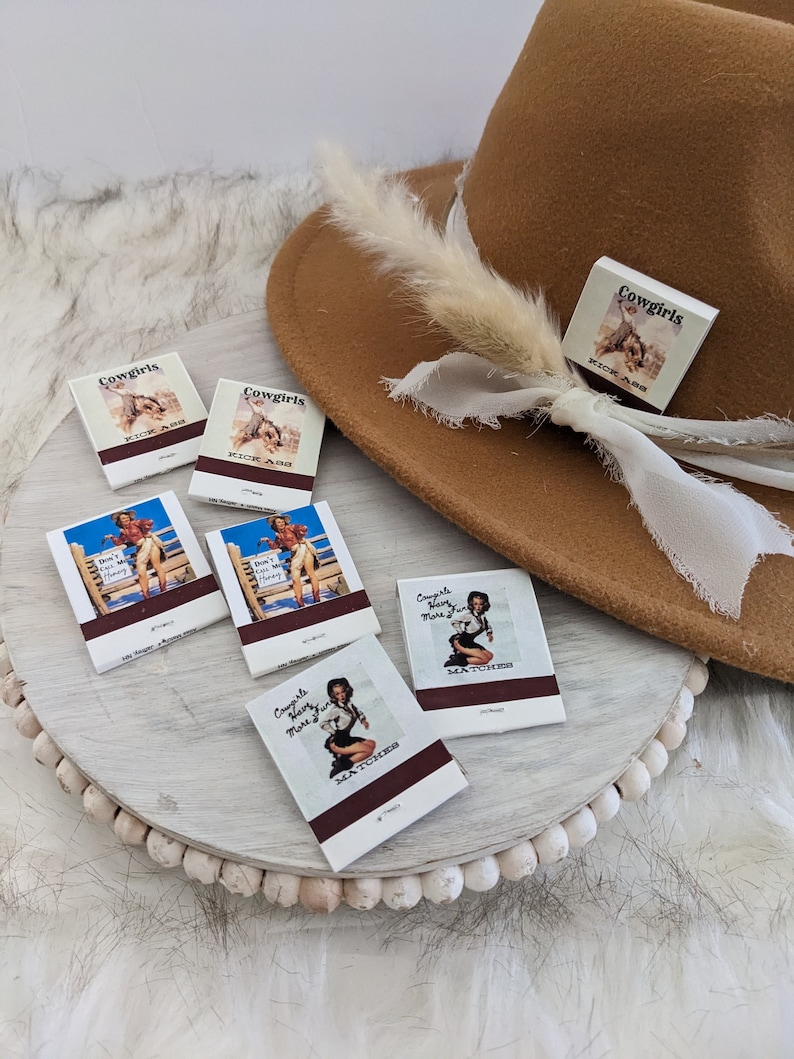 Pu&ograve; includere: Un set di sei fiammiferi con vari disegni che raffigurano cowgirl. I fiammiferi sono su una superficie di legno bianco con un cappello marrone e una piuma bianca sullo sfondo. I fiammiferi hanno il testo "Cowgirls Kick Ass" e "Cowgirls Have More Fun" stampato su di essi.